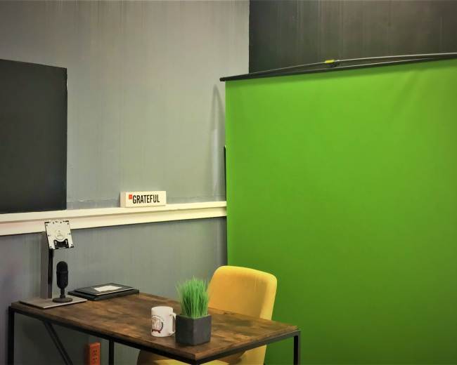 A small room with a green screen, a desk with a microphone, a plant, and a chair, along with a sign that reads "GRATEFUL."