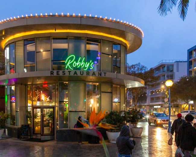 The image shows a modern circular restaurant with large glass windows, brightly lit signage, and people walking in a shopping area on a rainy evening.