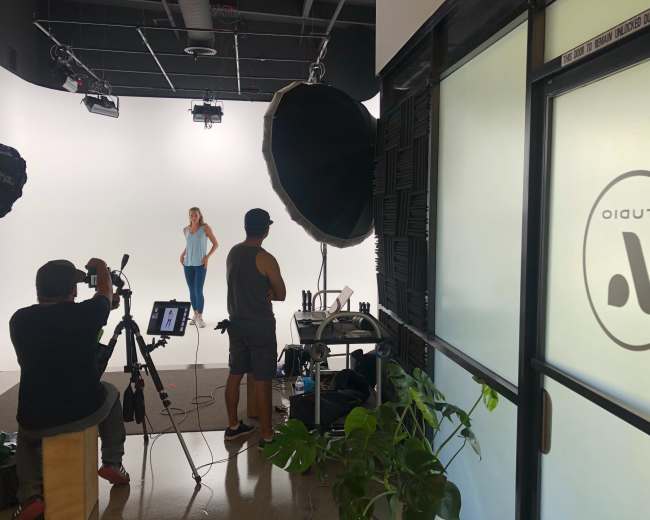 A woman poses in front of a white background while a camera crew sets up their equipment in a studio.