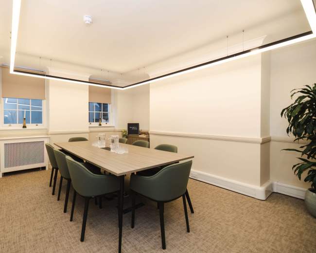 A well-lit meeting room with a rectangular table surrounded by six chairs, featuring a plant in one corner and a large window with blinds.