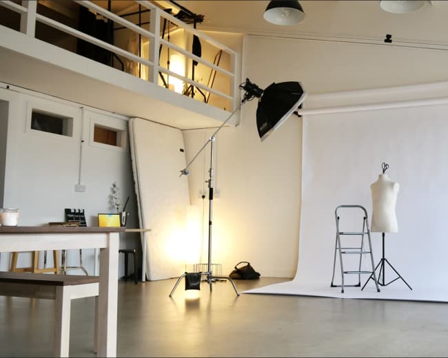 The image shows a photography studio with a white backdrop, a mannequin, lighting equipment, and a ladder set up for a photoshoot.