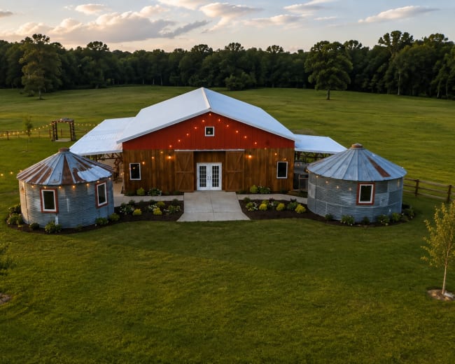 The image shows a barn-style building with a red wooden facade, surrounded by green grass and two silver silos on either side.