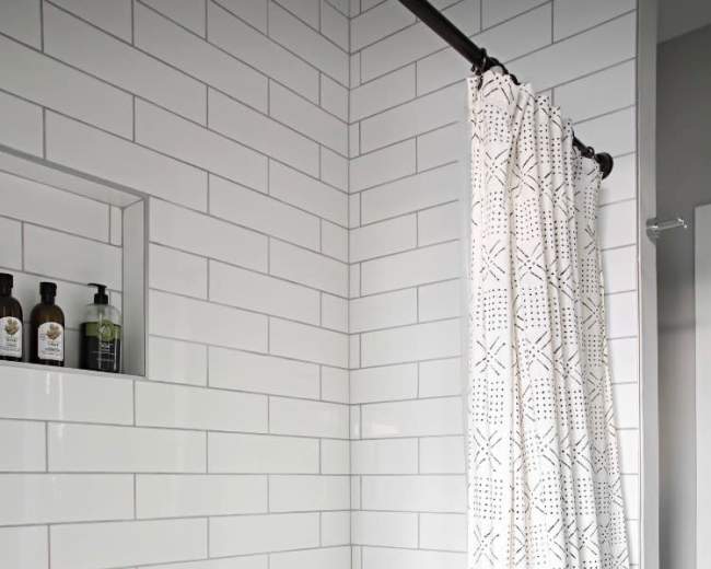 The image shows a white-tiled bathroom with a bathtub, a shower curtain with a geometric pattern, and a niche containing bottles on the wall.
