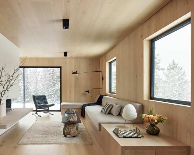 The interior features a minimalist living room with light wooden walls, large windows showcasing a snowy landscape, and a black lounge chair beside a small table adorned with flowers and a decorative item.