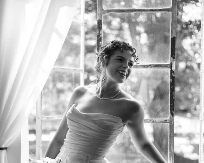 A bride in a strapless white dress stands by an open window, holding a bouquet of flowers and smiling while looking outside.