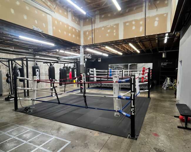 The image shows an indoor boxing gym featuring a competition boxing ring with red and blue corners, surrounded by training bags and a spacious floor.