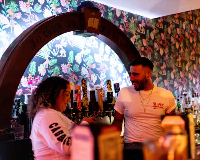 A bartender converses with a staff member in a bar featuring a vibrant, floral wallpaper and a variety of drink taps.
