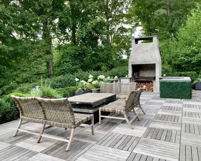 The image shows a landscaped outdoor patio area with a seating arrangement of woven chairs and a central table near a stone barbecue grill surrounded by greenery.