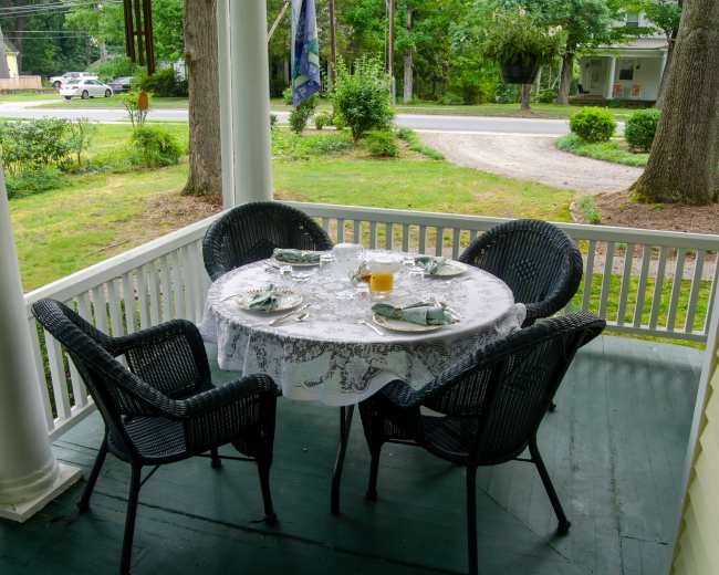 A round table set with dishes and drinks is situated on a porch surrounded by greenery.