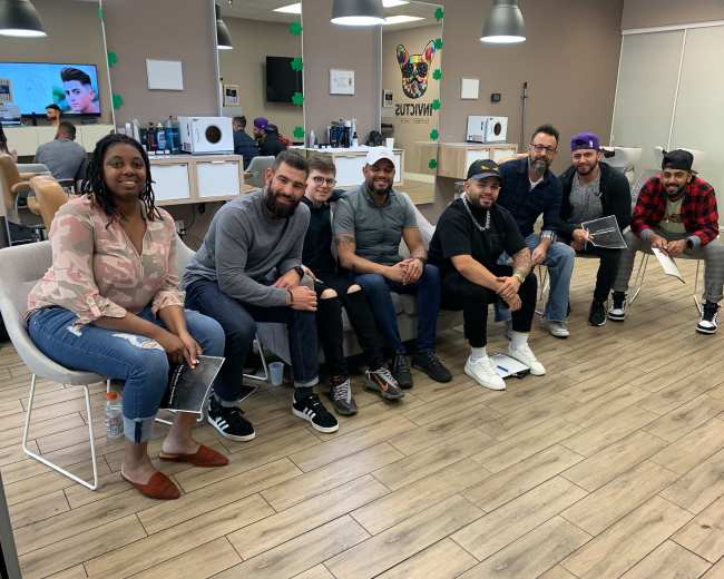 A group of eight people is seated in a barber shop, with grooming stations visible in the background.