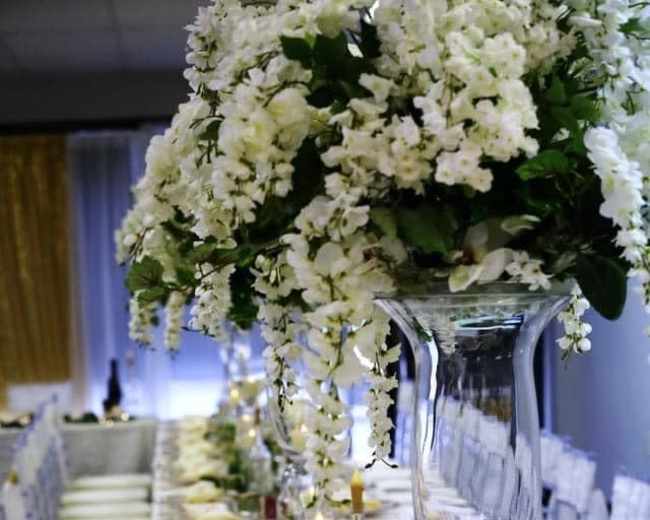 The image shows a long dining table elegantly set with white chairs, featuring decorative floral arrangements and candles in tall glass vases.