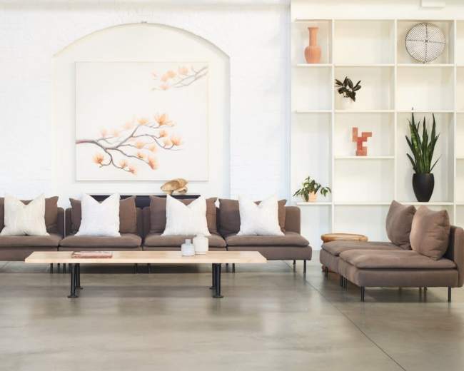 A modern living room features a long, brown sectional sofa with white pillows, a wooden coffee table, potted plants, and minimalist decor against a white brick wall.