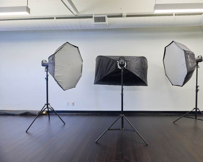 The image features three studio light setups on stands arranged in an empty room with dark wooden flooring.