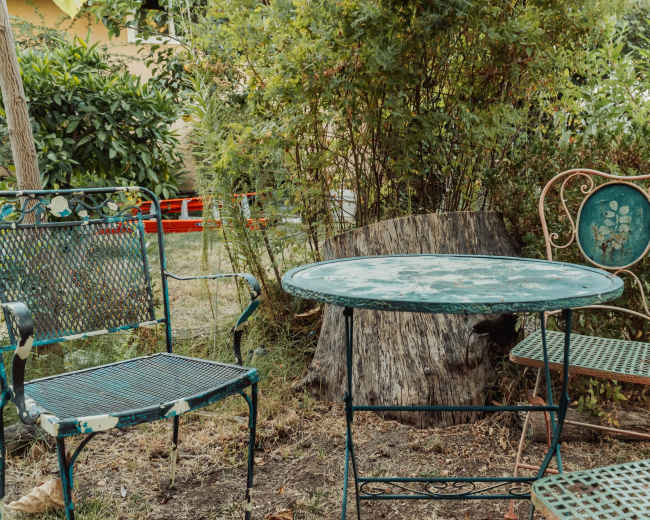 A small outdoor seating area features a round table surrounded by metal chairs, set against a backdrop of greenery and a tree stump.