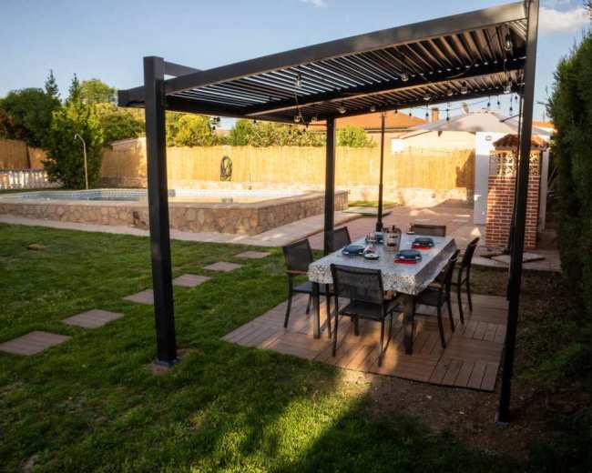 The image shows a backyard scene with a modern pergola over a dining table and chairs next to a swimming pool, surrounded by greenery and a fence.