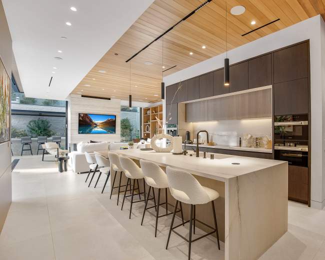 A modern kitchen features a large island with bar seating, sleek cabinetry, and a view of an outdoor space with a television display.