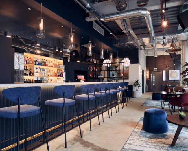 The image shows a modern bar area with a long counter, several stylish high stools, and a mix of seating arrangements including lounge chairs and tables.