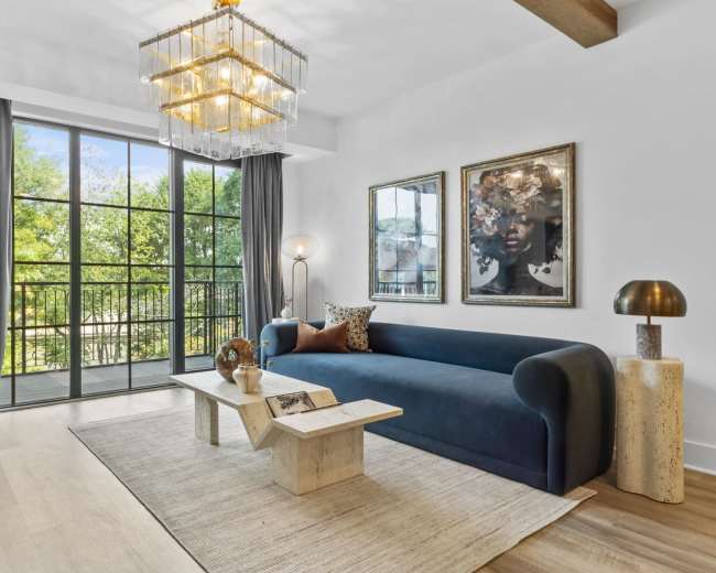 The image shows a modern living room featuring a blue sofa, a coffee table made of stone, and large windows with views of greenery outside.