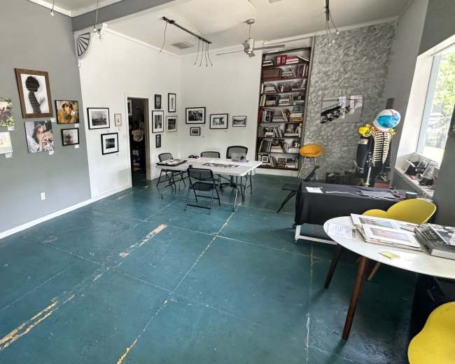 The image shows an art gallery with framed photographs on the walls, tables set up in the center, and bookshelves in the background.