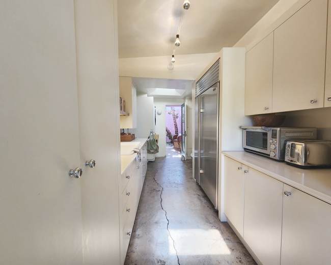 A narrow kitchen corridor featuring white cabinetry, stainless steel appliances, and visible natural light leading to an outdoor area.