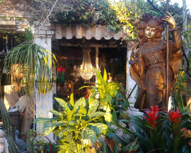 A large statue of a figure stands beside a lush garden filled with various plants, while a chandelier is visible in the background through an open doorway.