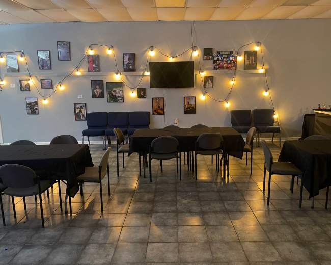 The image shows an empty event space arranged with black tablecloths, chairs, and framed photographs on the walls, illuminated by warm string lights.