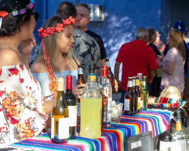 Two women, one wearing a floral dress and a flower crown, are standing behind a table adorned with colorful fabric, surrounded by various bottles of alcohol and a crowd in the background.