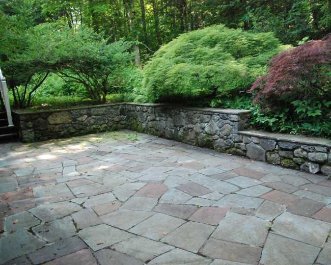 A stone patio is surrounded by lush greenery and shrubbery along a low stone wall.