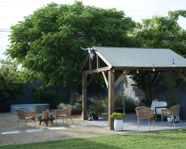 A wooden pergola with a metal roof, surrounded by a landscaped garden featuring a fire pit, outdoor seating, and a large tree.
