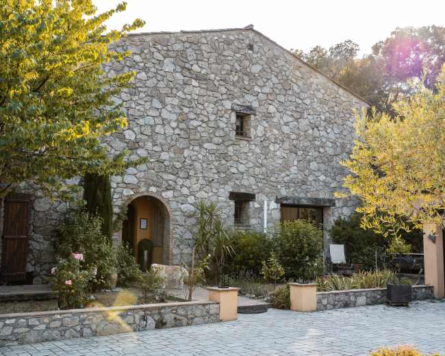 A rustic stone house with a sloped roof is surrounded by greenery and flowering plants in a peaceful setting.