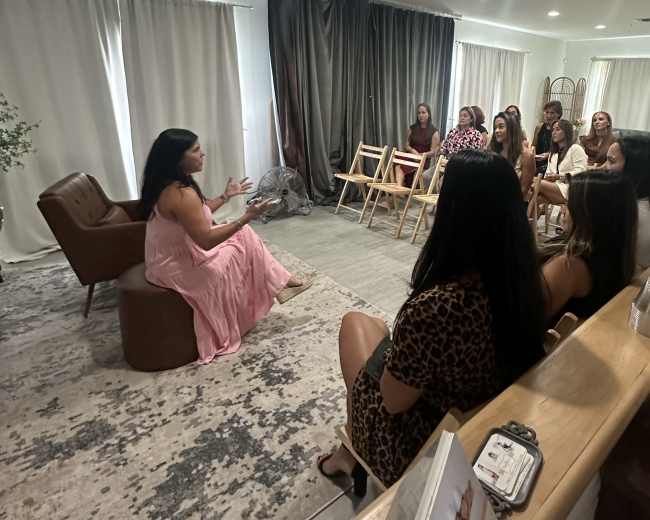A woman in a pink dress speaks to a group of seated individuals in a softly lit room with neutral-colored curtains and a rug.