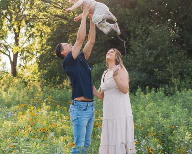 A man lifts a child above his head while a woman smiles in a field of wildflowers under a bright sun.