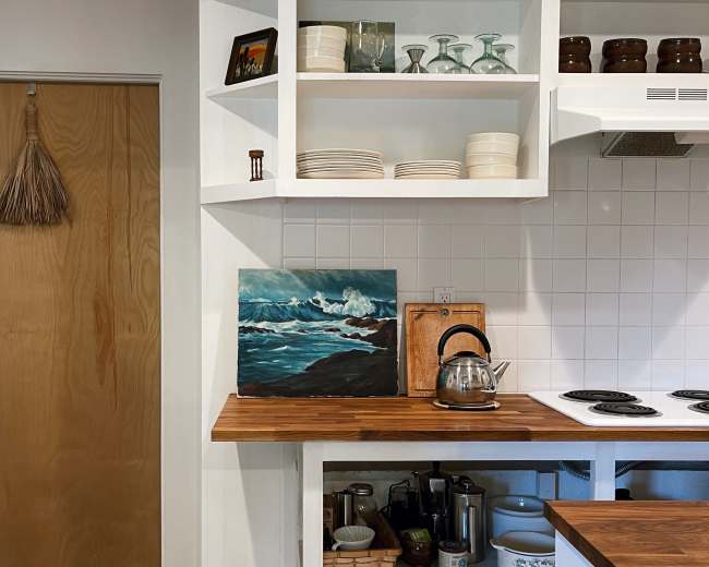 A kitchen scene features a wooden countertop with a kettle and an artwork, alongside shelves displaying glassware and dinnerware, next to a closed door.