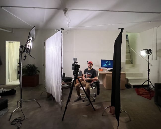 A person sits on a chair in a room set up for video recording, surrounded by lighting equipment and backdrops.
