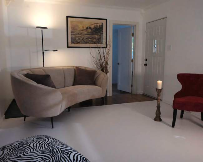 The image features a cozy living room with a beige curved sofa, a red accent chair, a candle on a stand, and a large framed landscape picture on the wall.