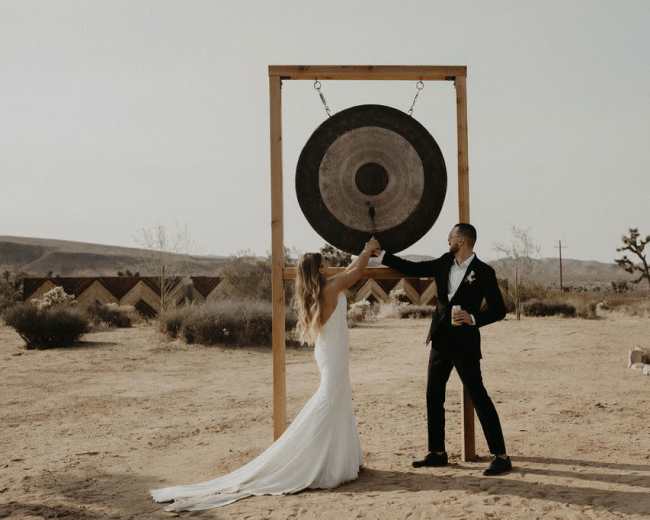 A couple in formal attire strikes a large gong in a desert setting, with desert plants and mountains in the background.