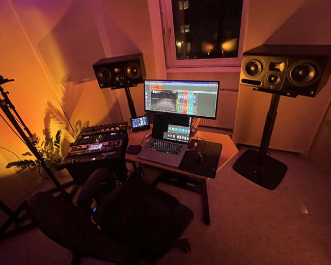 A music production setup features a computer monitor, audio interface, and two studio monitors in a room with low ambient lighting.