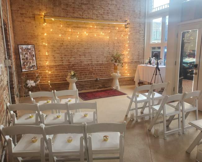 A small wedding ceremony setting features white folding chairs arranged in front of a decorated backdrop with string lights, flanked by a table and floral arrangements.