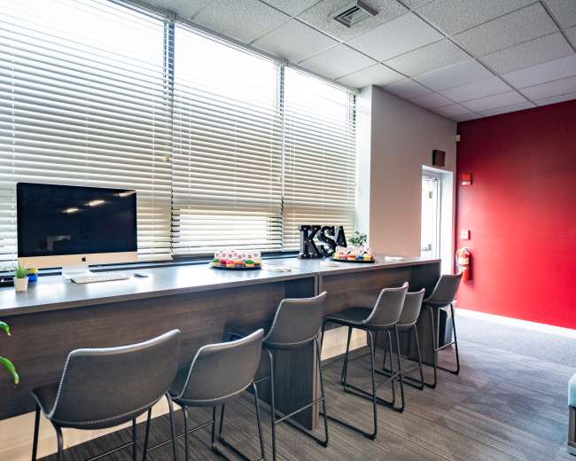 The image shows a modern office space with a long counter featuring several high-backed stools, a computer, and decorative items against a backdrop of large windows and a red accent wall.