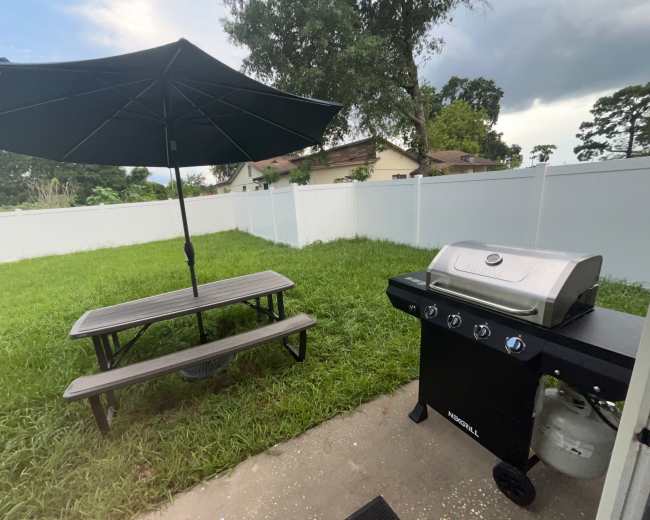 The image shows a barbecue grill beside a picnic table with an umbrella in a yard surrounded by a white fence and tall grass.