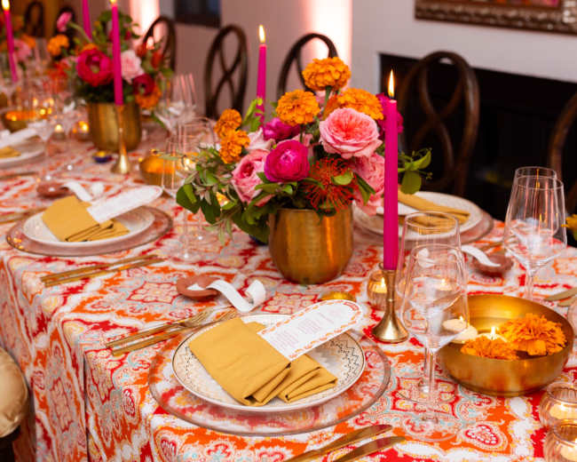A festively set dining table features vibrant floral arrangements, gold accents, and colorful tableware.