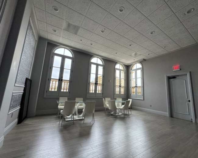 The image shows a spacious room with large arched windows, wooden flooring, and modern white furniture, including tables and chairs.