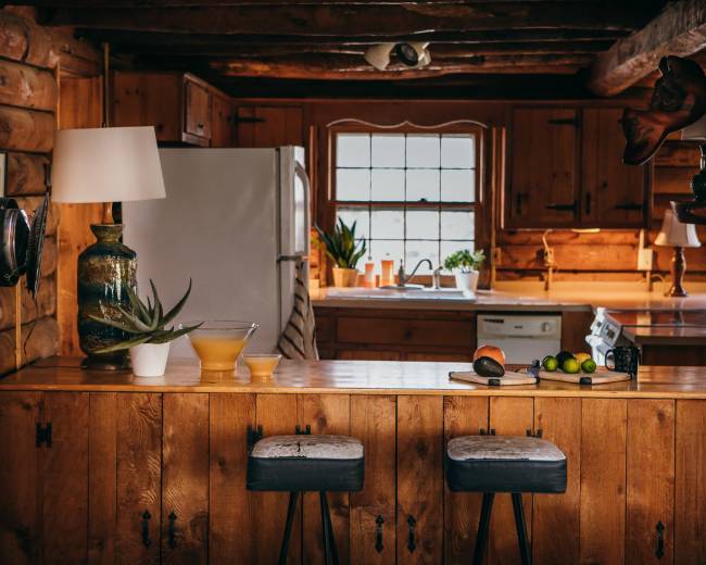 A rustic kitchen features wooden cabinetry, a bar with two stools, and natural light streaming through a window.
