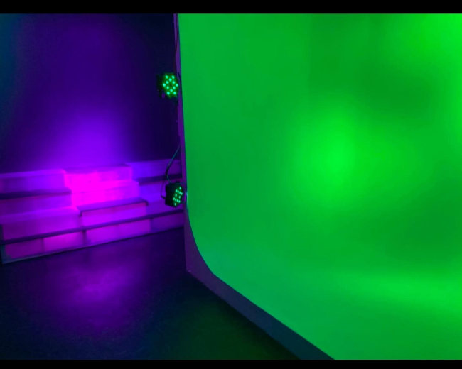 The image shows a studio space with a green backdrop and purple lights illuminating the area.