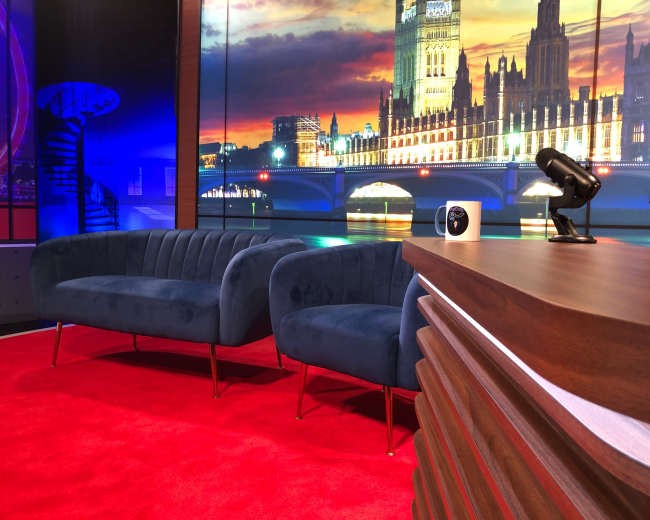 The image features a modern television studio setup with blue sofas, a wooden desk, and a backdrop of a lit-up city skyline.