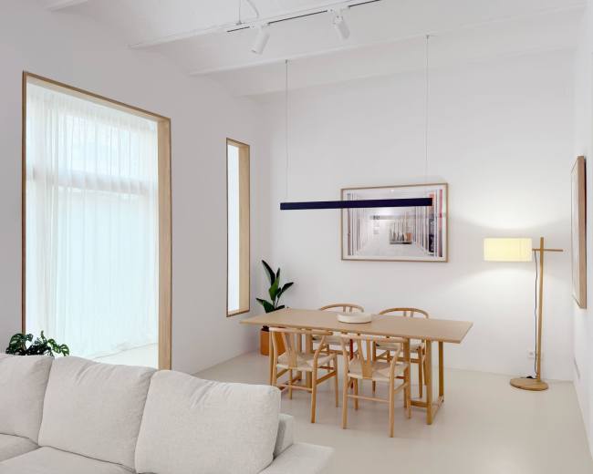 The image shows a minimalist dining area with a wooden table surrounded by chairs, illuminated by a modern light fixture, and featuring large windows with sheer curtains.