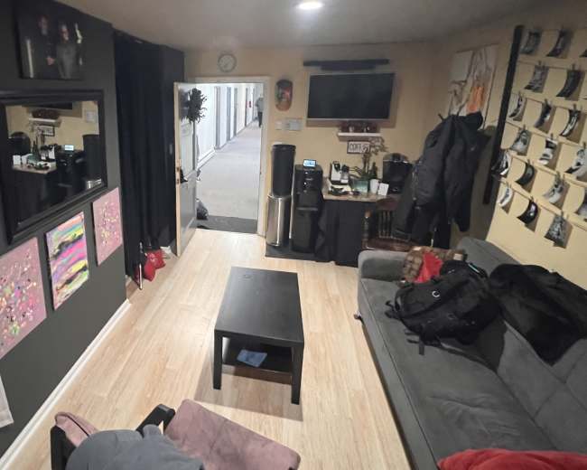 A casual living space with a couch, a coffee table, and various items like bags and a coffee machine, along with wall decorations.