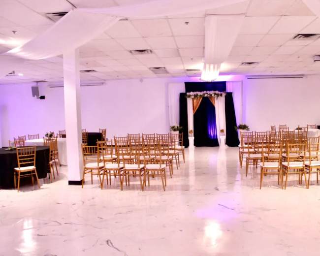 A spacious event venue features rows of tables with gold chairs arranged around a central decorated backdrop.
