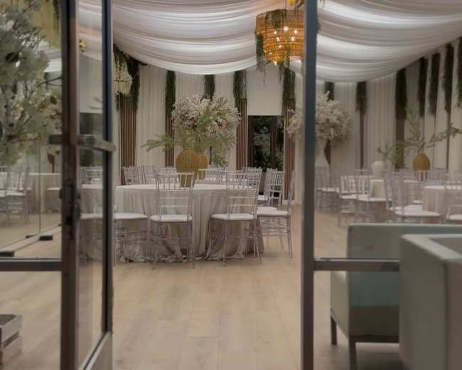 The image shows a well-decorated indoor event space with draped ceilings, tables set for an occasion, and floral arrangements throughout.