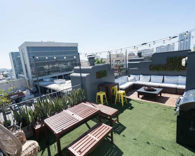 The rooftop features wooden furniture, green plants, and a seating area with a grill against a backdrop of urban buildings and clear blue skies.